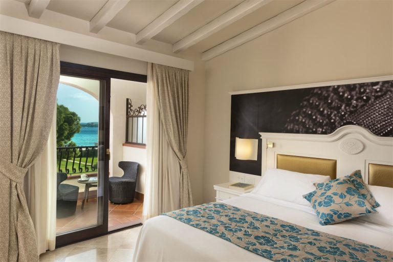un letto matrimoniale con una grande testiera lavorata in legno, si affaccia su una veranda con vista sul mare e sul giardino