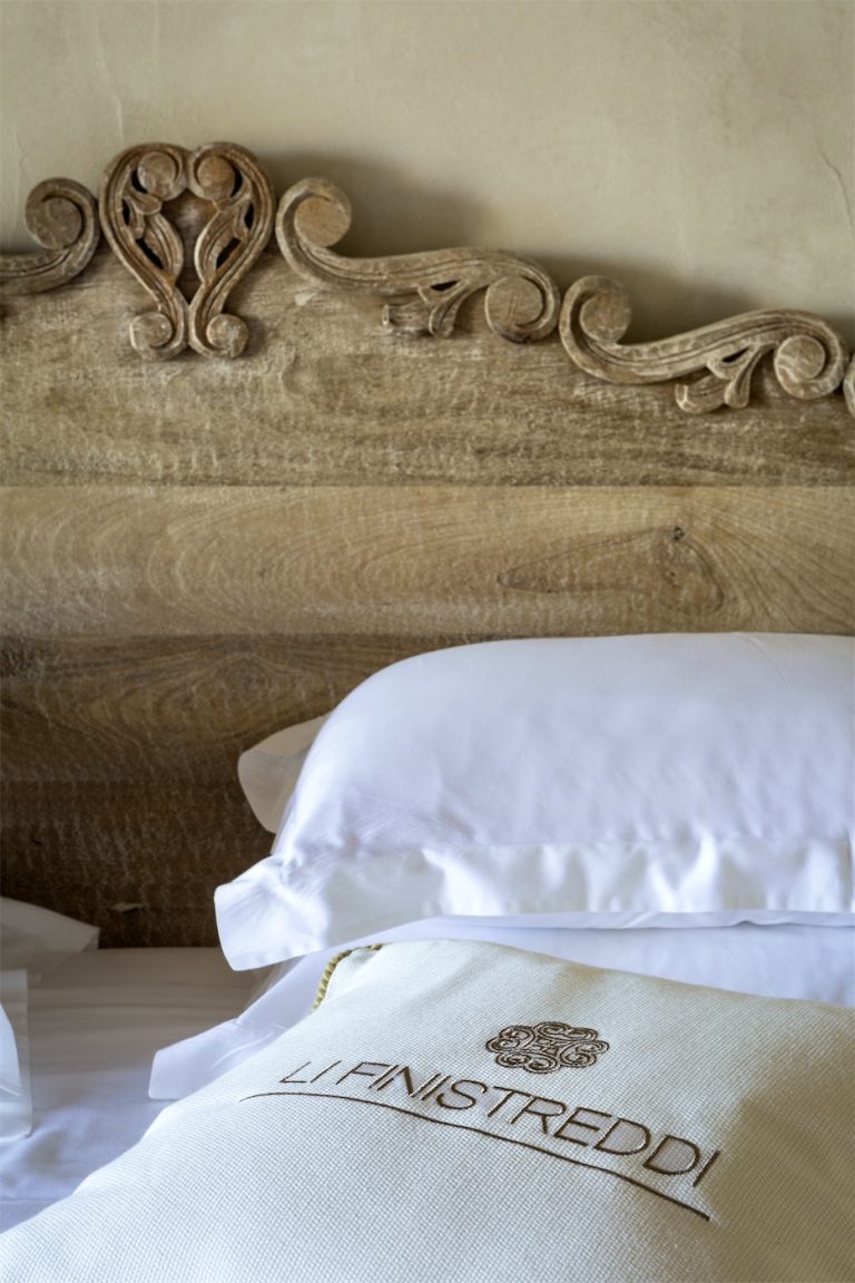 particolare della testata in legno di un letto matrimoniale, finemente intagliata, con un cuscino su cui sono ricamati nome e logo dell'hotel Li Finistreddi