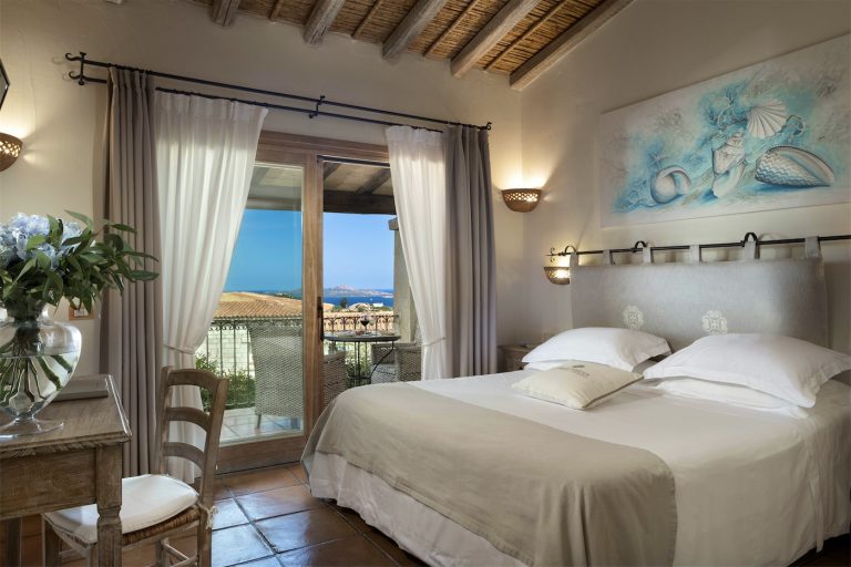 letto matrimoniale con una testiera dai colori neutri. Decorazioni marine sulla parete ed una vetrata che si apre su una terrazza con vista sul mare