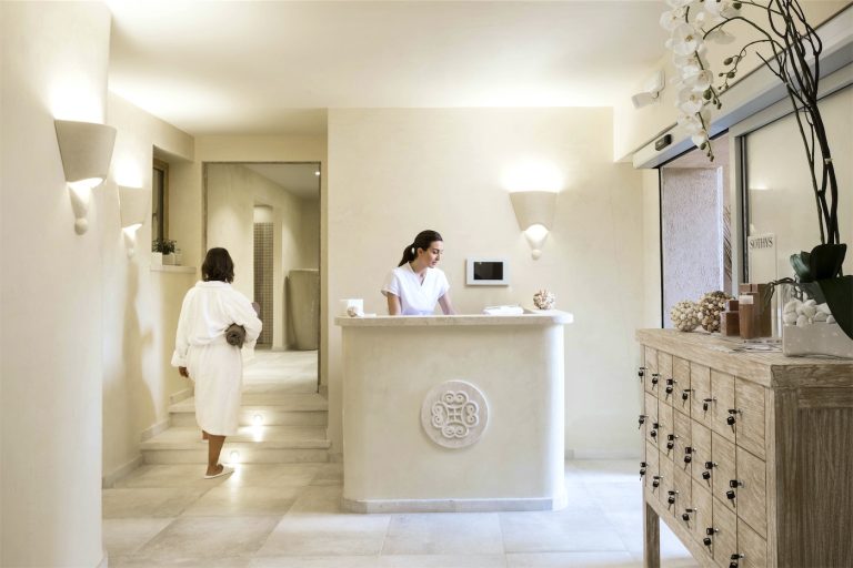 Una operatrice al banco reception della spa dell'hotel, vicino ad una cliente in accappatoio ed un mobile con degli sportelli porta oggetti