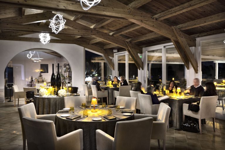 una grande sala illuminata per la sera, con vari tavoli, clienti che mangiano ed un soffitto in legno con grosse travi scure